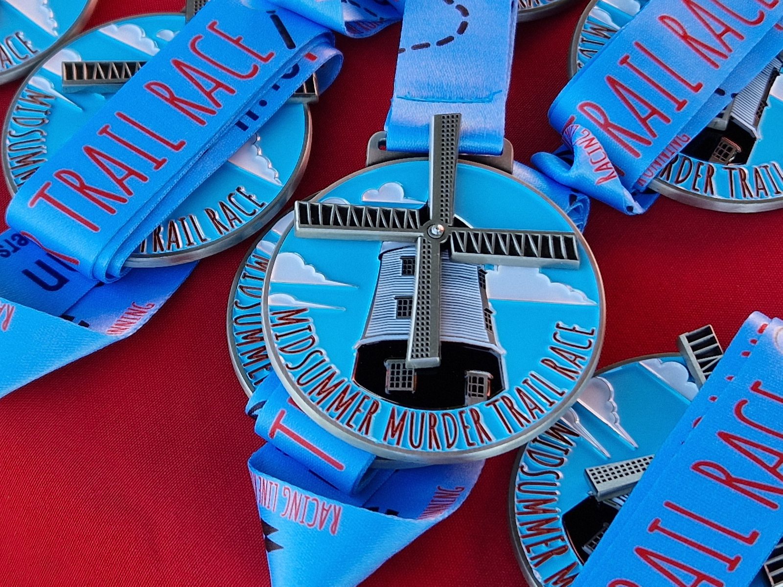 A close-up of several blue and silver medals for the "Midsummer Murder Trail Race." The medals depict a windmill against a blue sky with puffy clouds. The blue lanyards attached to the medals are printed with the event name "Midsummer Murder Trail Race.