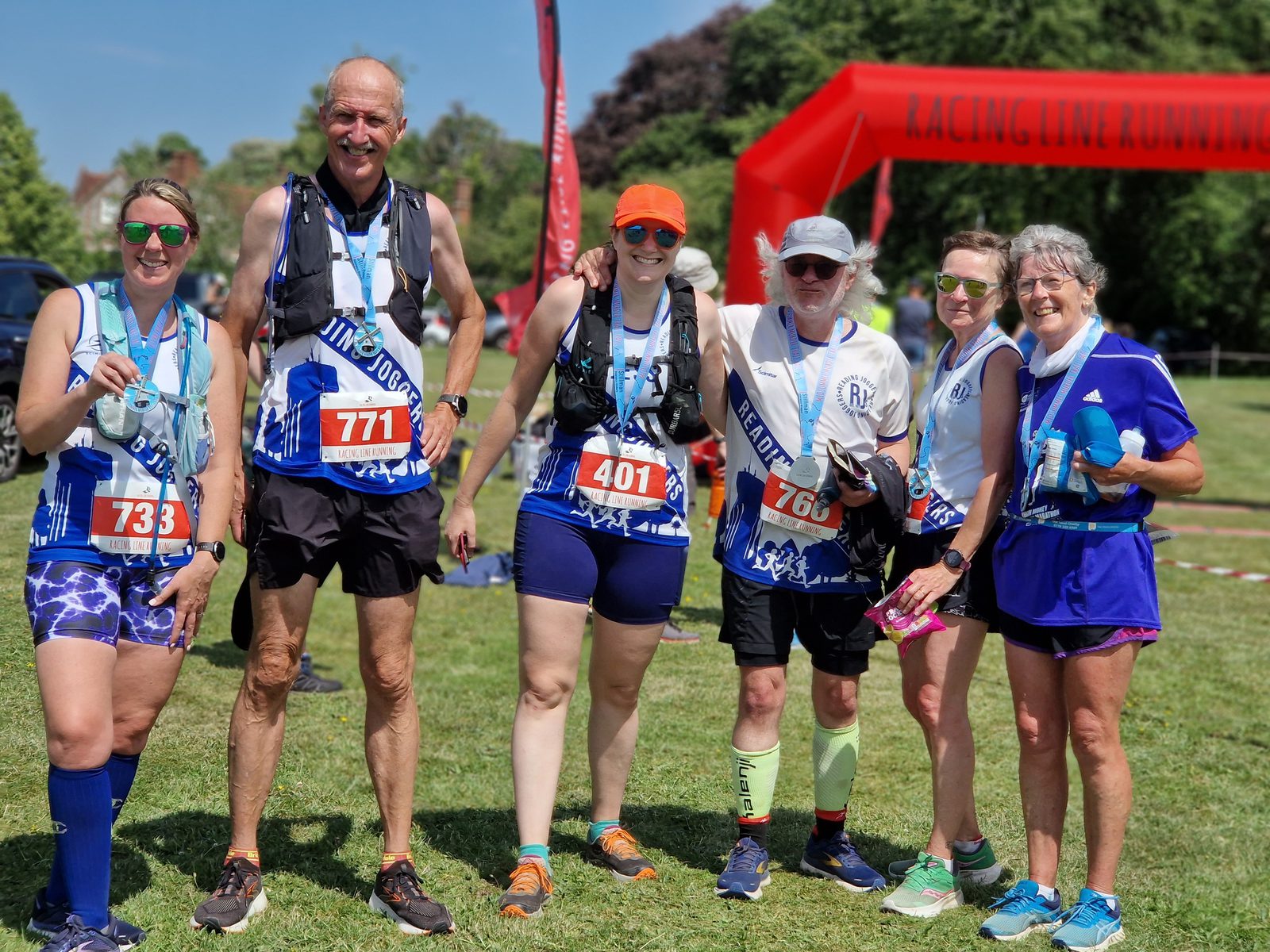 A group of six runners pose for a photo after a race. They are wearing bib numbers and medals, with four holding water bottles. They stand on grass with a red arch and trees in the background, all smiling and wearing athletic gear, including shorts and tank tops.