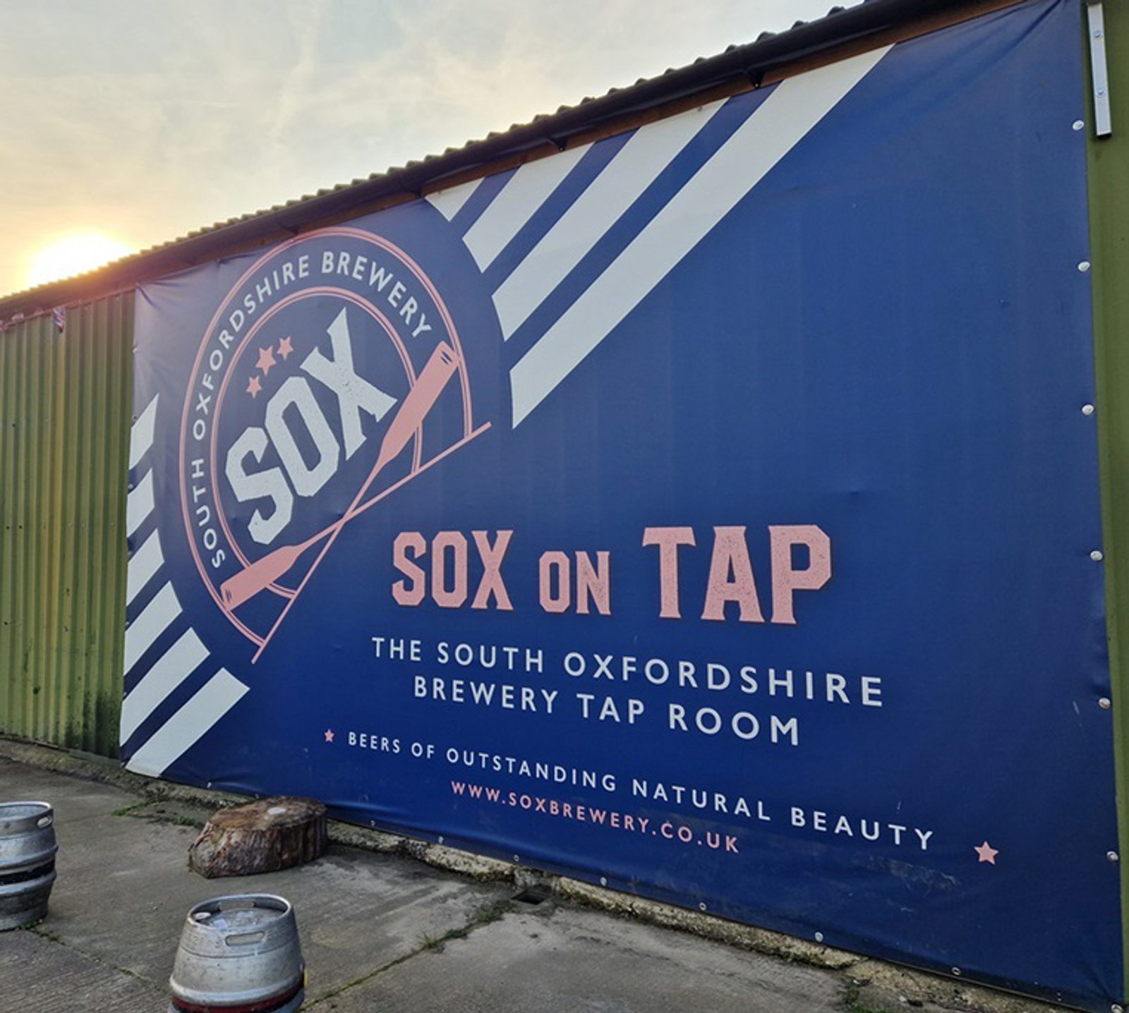An outdoor billboard reads "SOX on Tap. The South Oxfordshire Brewery Tap Room. Beers of Outstanding Natural Beauty." The background is blue with diagonal white stripes and the brewery logo in white and pink. The bottom of the sign lists the website: www.soxbrewery.co.uk.
