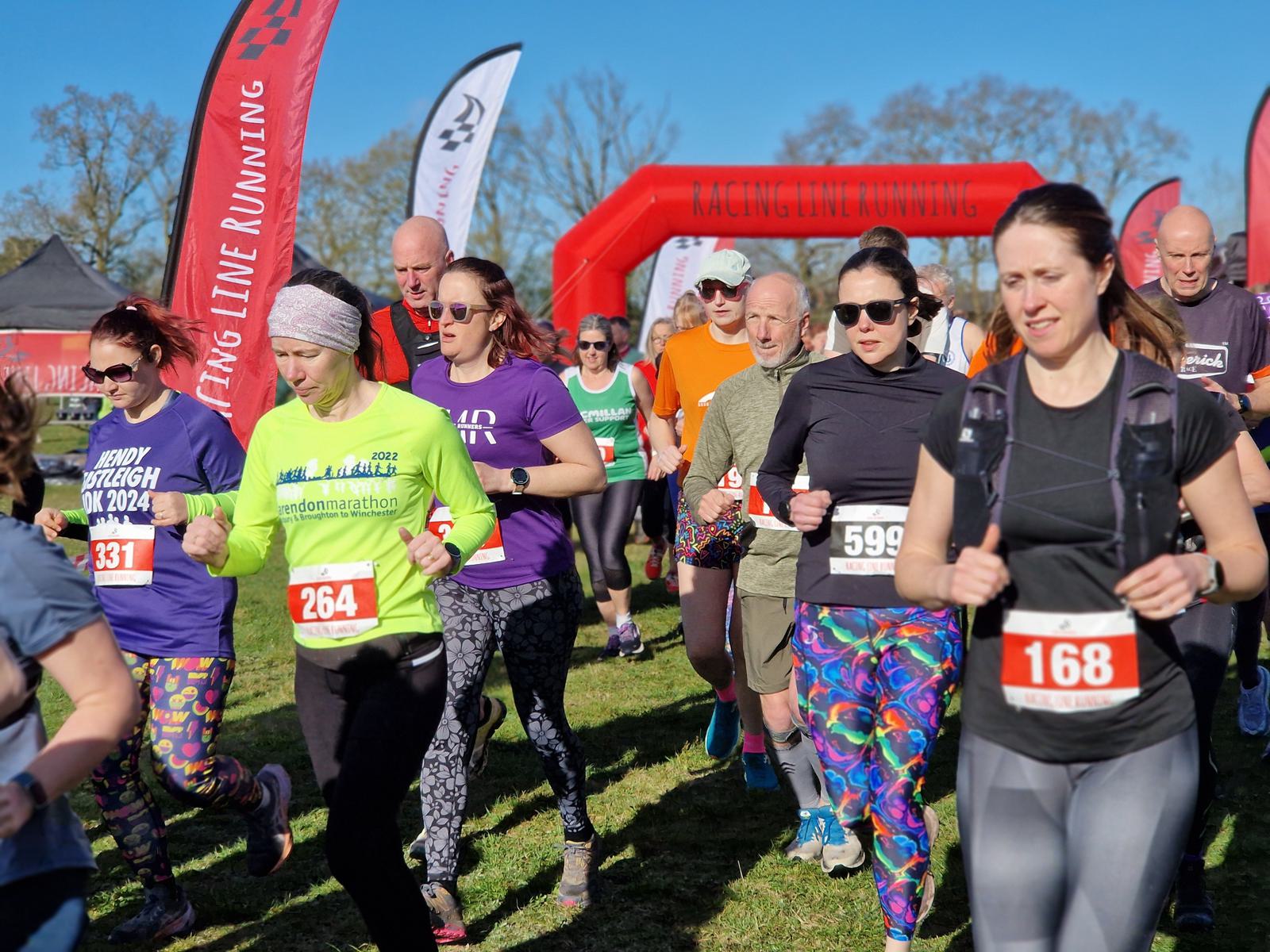 Runners participating in a marathon on a sunny day, wearing colorful athletic gear with bib numbers. Red banners and an archway from "Racing Line Running" are visible in the background, along with a clear blue sky.
