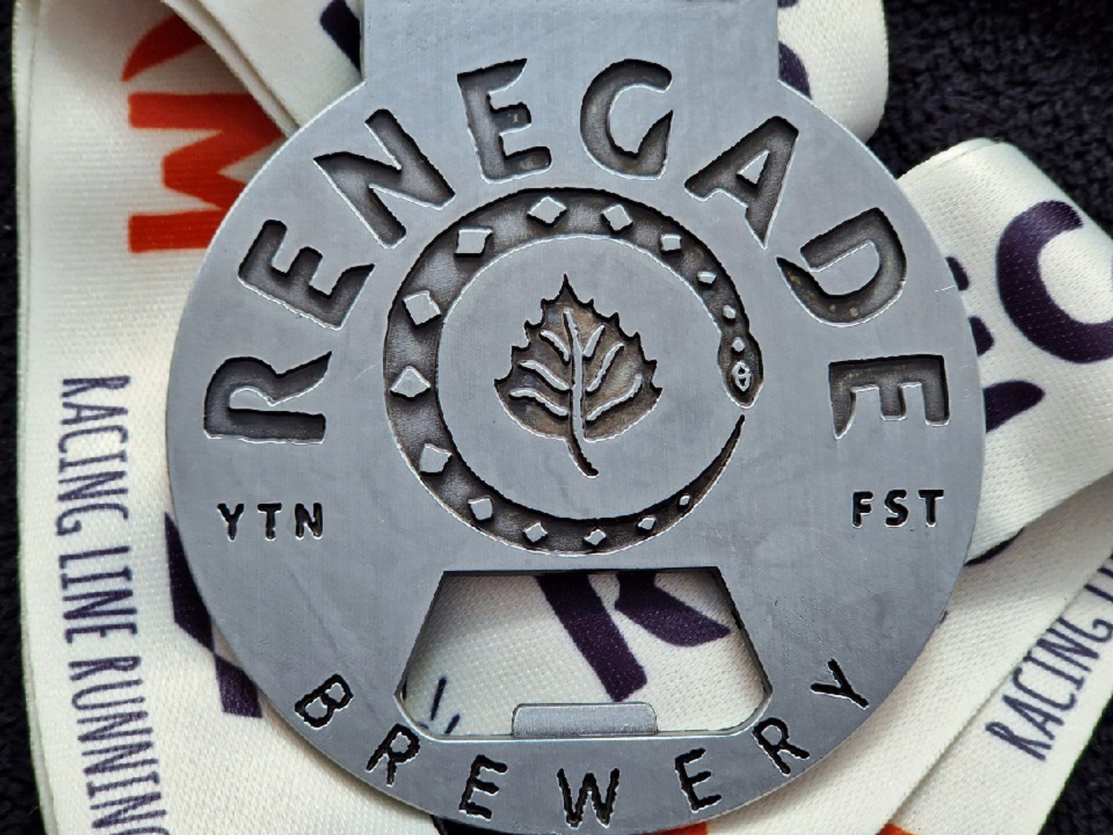 Close-up of a metallic Renegade Brewery medal featuring a central circular design with a leaf and snake. The ribbon has partial text that reads "RACING LINE RUNNING.