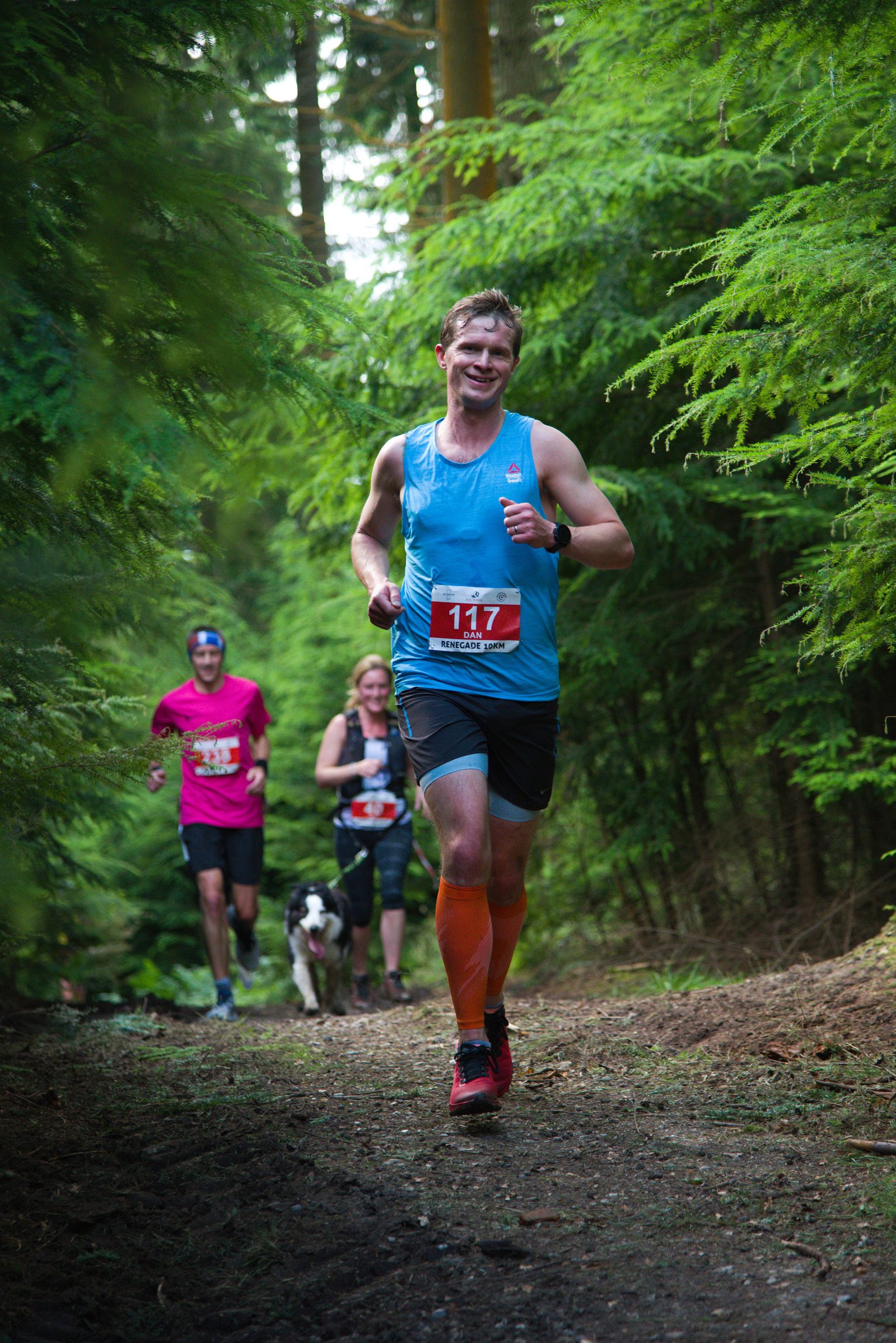 A smiling runner in a blue tank top and number 117 bib jogs through a forest trail. Behind him, another runner, a woman, and a dog are also on the path, surrounded by lush green trees. It's a vibrant outdoor scene.