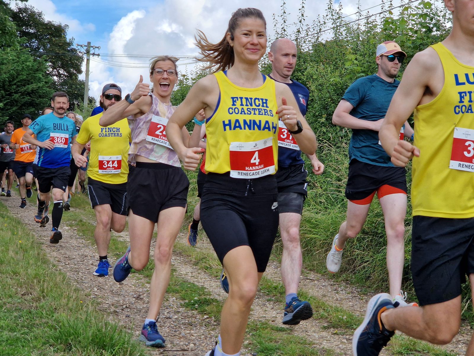 A group of runners in athletic gear running on a dirt path during a race. A woman in a yellow "FINCH COASTERS" tank top leads the group, while others follow closely, some smiling and giving a thumbs up. The weather is clear and sunny.