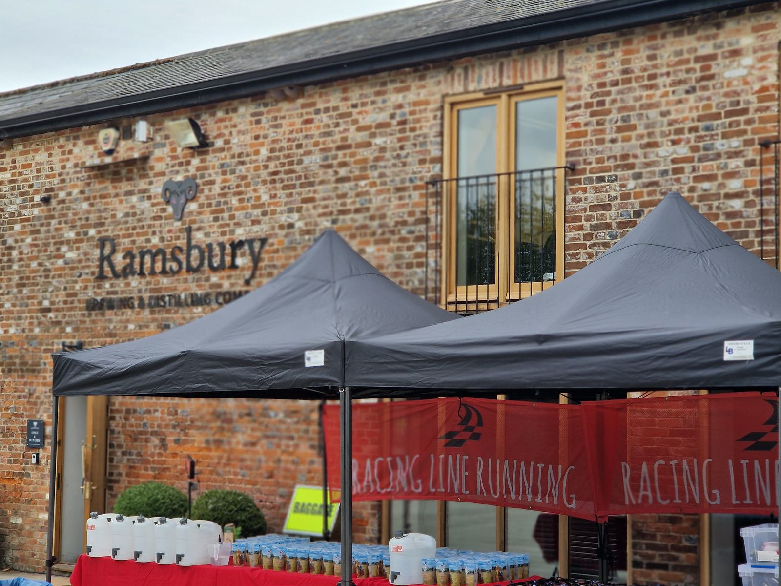 Outdoor scene of a brick building with "Ramsbury Brewing & Distilling Co." on the wall. Two black tents with a red banner reading "Racing Line Running" underneath. Tables hold jugs and bottles.
