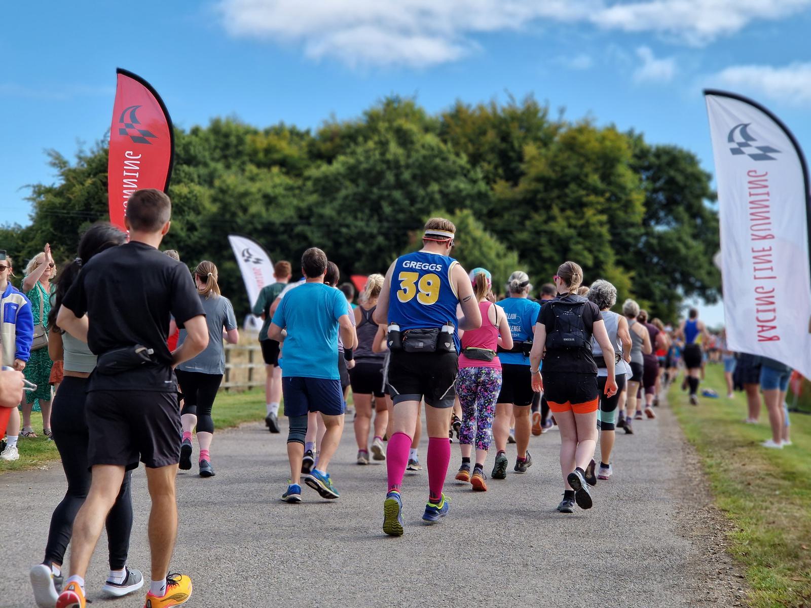 Runners participate in an outdoor race on a sunny day, heading down a paved path lined with trees. Participants wear colorful attire, and race banners are visible along the route. One runner prominently wears a jersey with "Greggs 39" on the back.