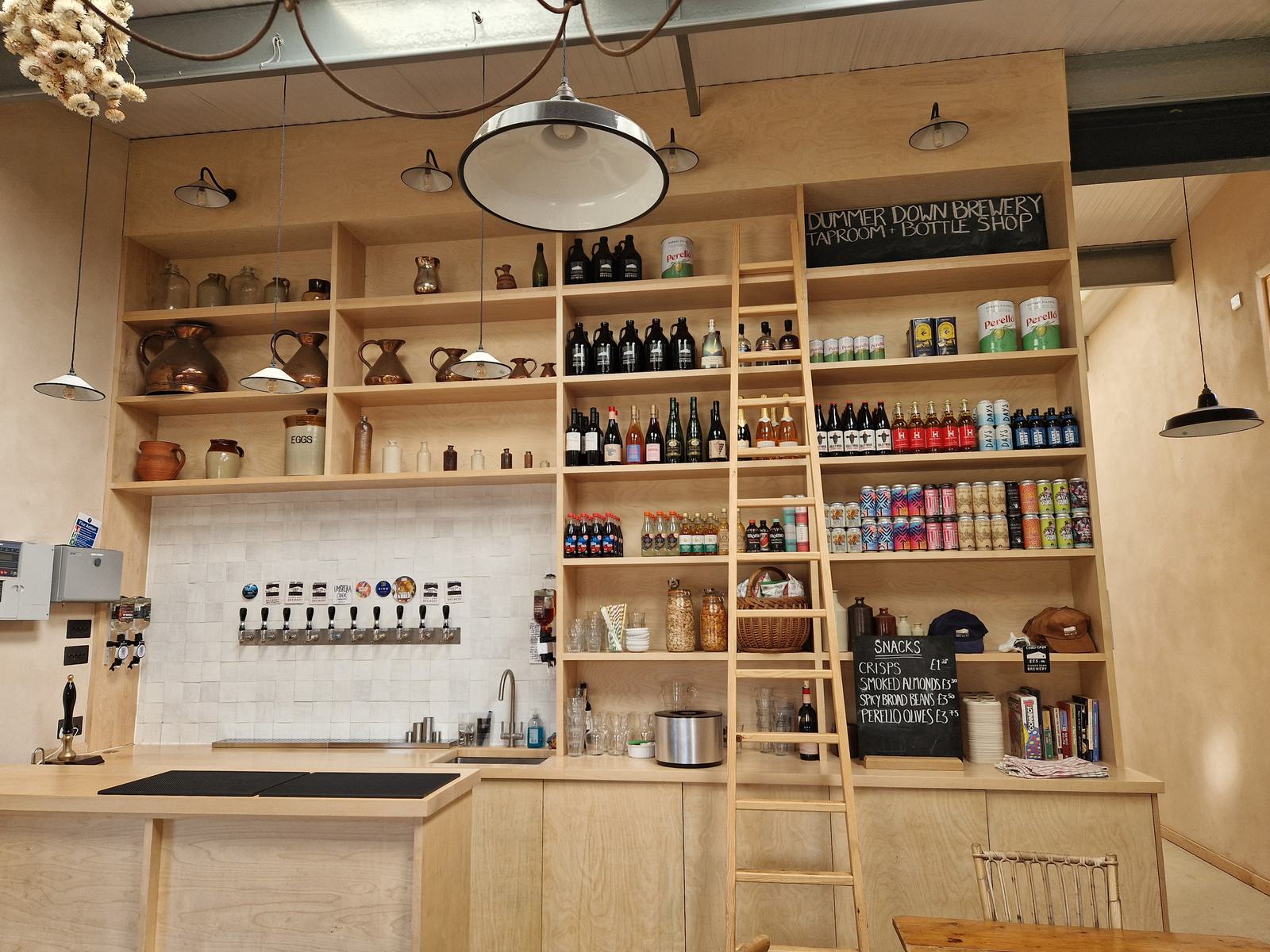 A cozy brewery interior with a wooden bar, taps, and shelves stocked with various bottles and cans. A ladder leans against the wall for reaching the high shelves. Warm lighting and wooden accents create a welcoming atmosphere.