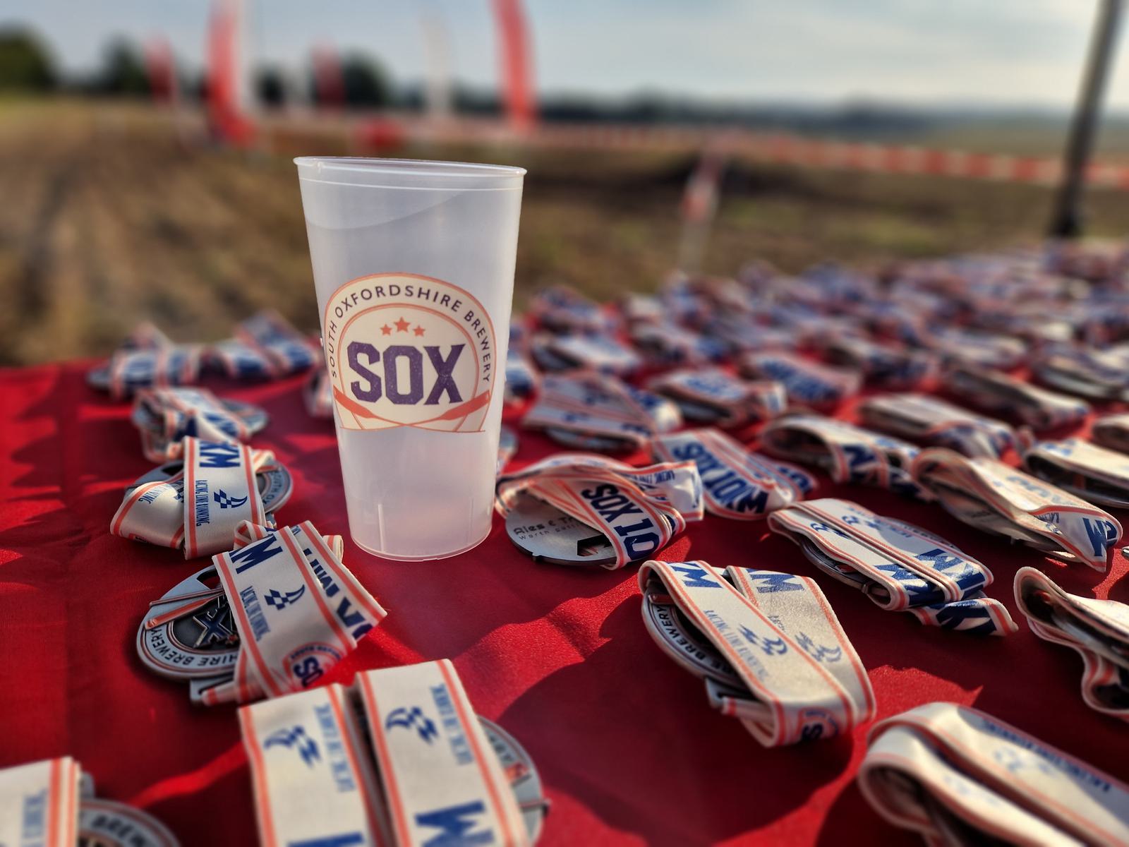A plastic cup with "SOX" printed on it stands on a red table covered with numerous medals. The background features an outdoor field with blurred red and white decorations.
