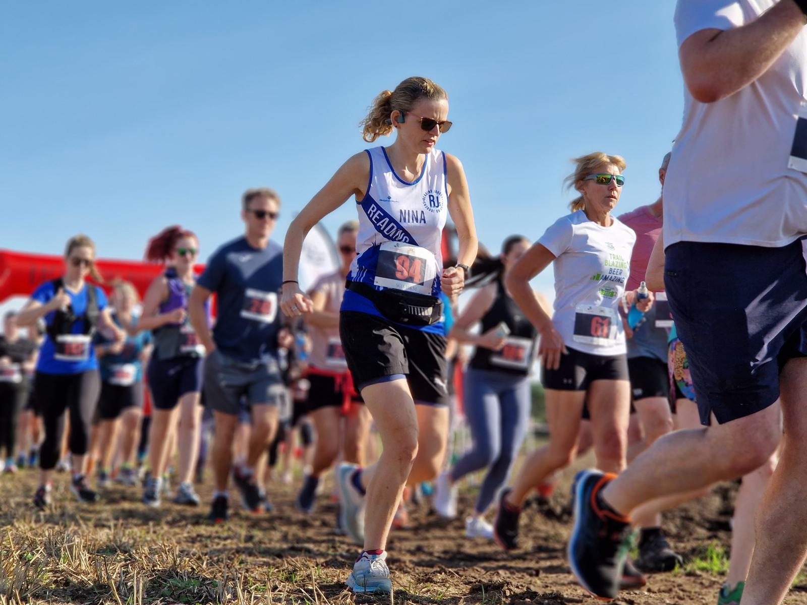 A group of runners participating in an outdoor race on a sunny day. A woman in the foreground wears a white and blue tank top and shorts, with a race number pinned to her outfit. Other runners surround her, all in motion.