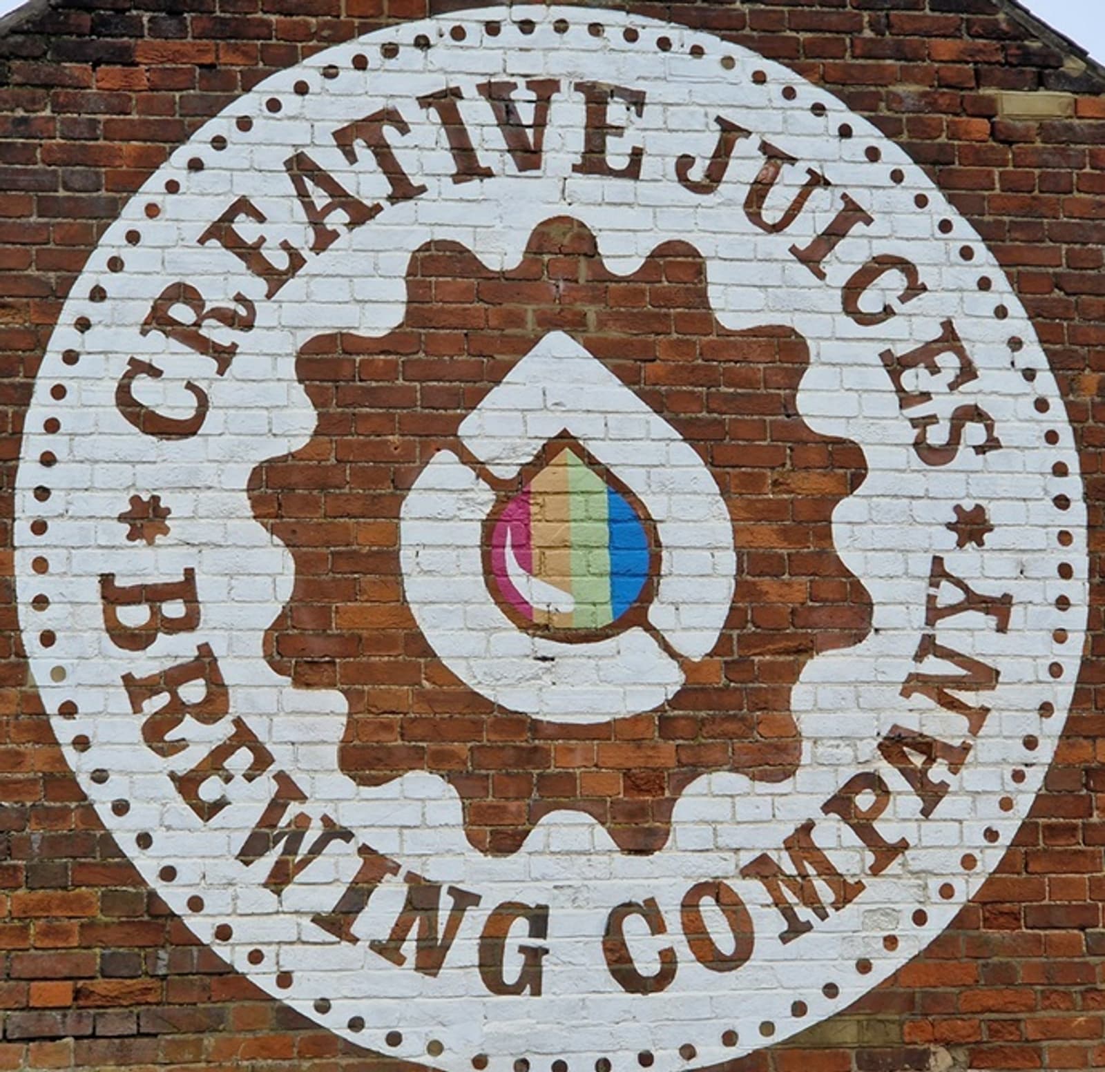 A circular mural on a brick wall displays the words "CREATIVE JUICES BREWING COMPANY" around a gear and droplet logo, featuring a rainbow-striped center.