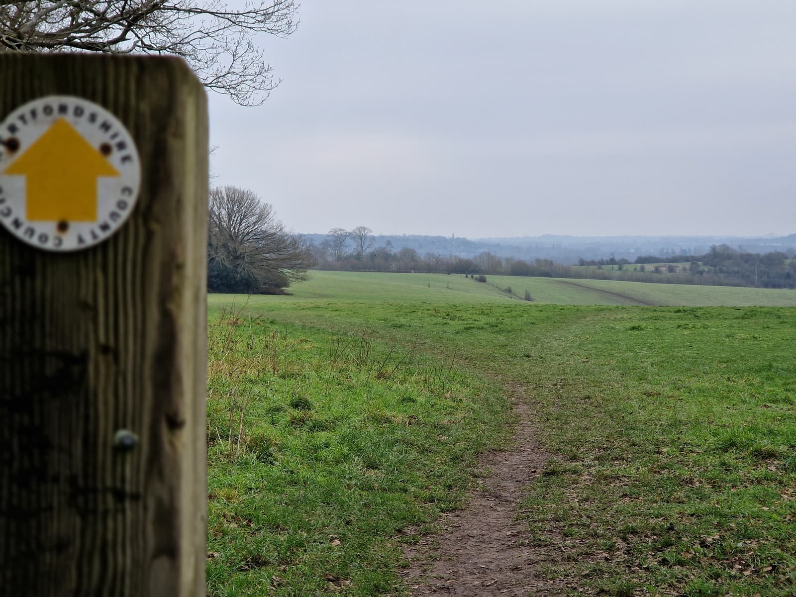 A dirt footpath runs through a grassy field toward distant hills under a cloudy sky. In the foreground, a wooden post displays a yellow arrow, marking the Oxfordshire County boundary path.