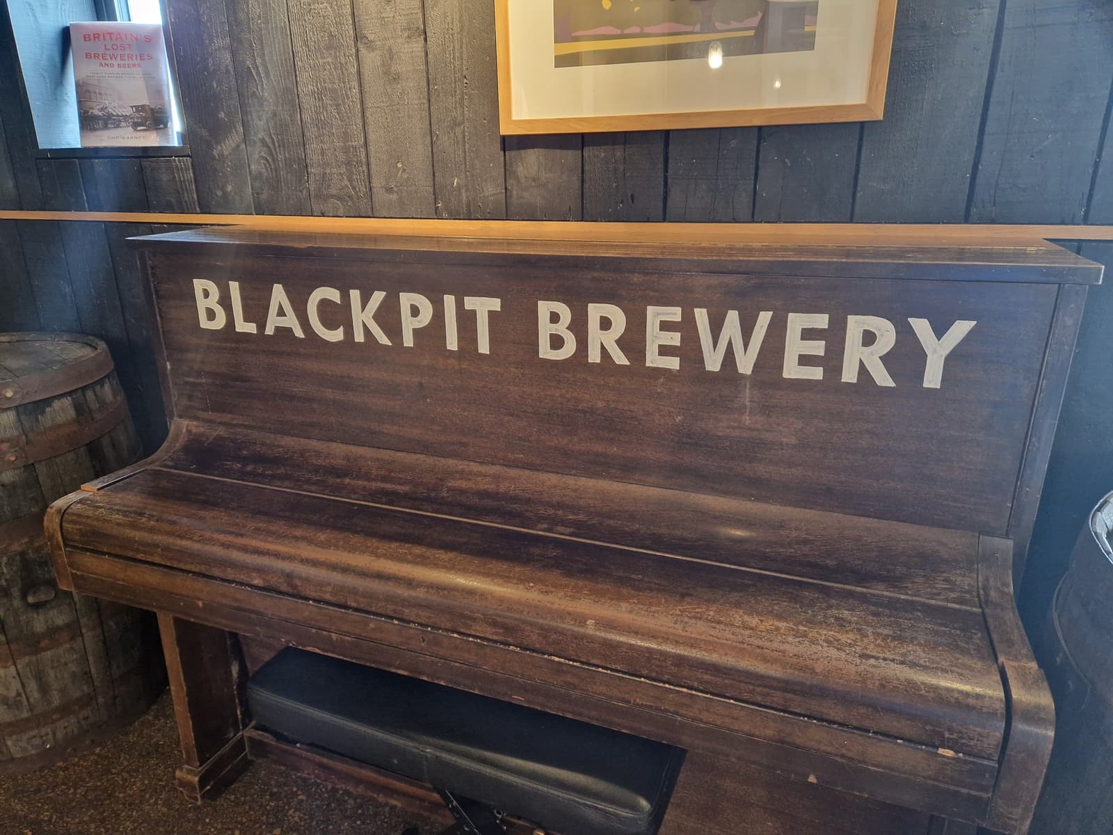 A wooden upright piano with "Blackpit Brewery" painted on the front, placed against a dark wooden wall inside a rustic setting. A barrel and framed pictures are partially visible nearby.