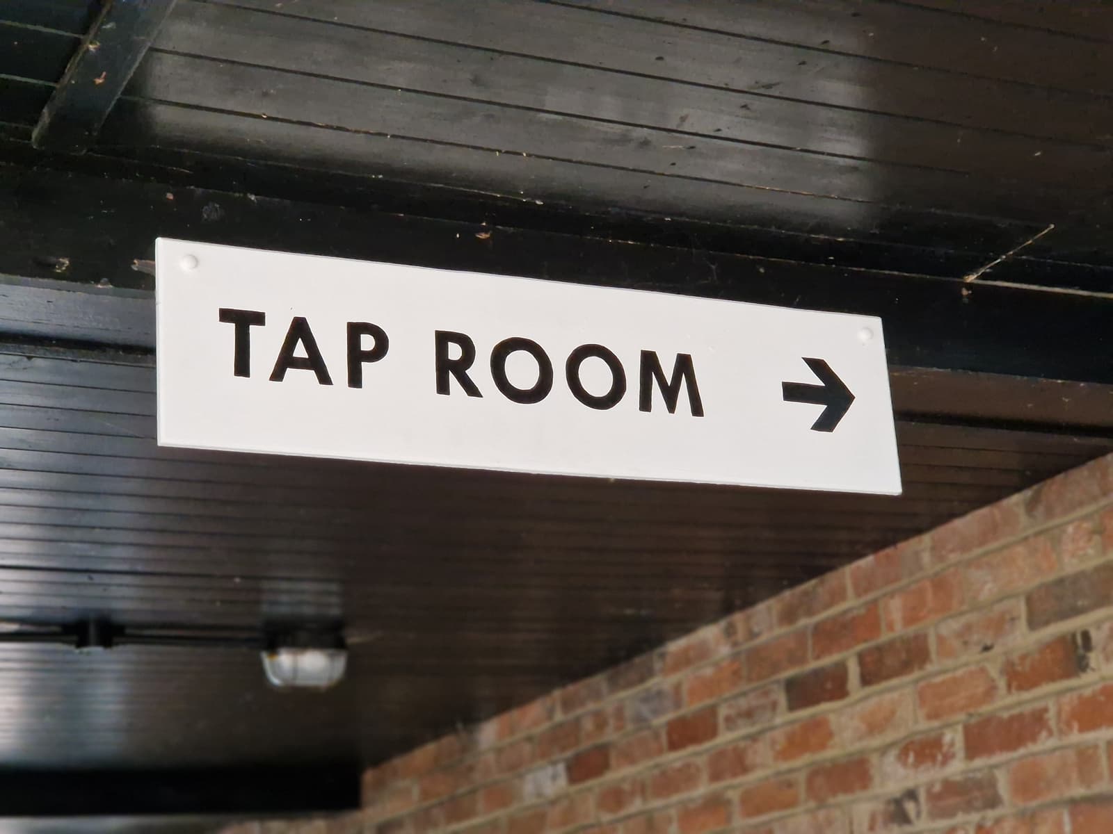 A white sign with black text reading "TAP ROOM" and a right-pointing arrow is mounted on a dark wooden ceiling above a brick wall.