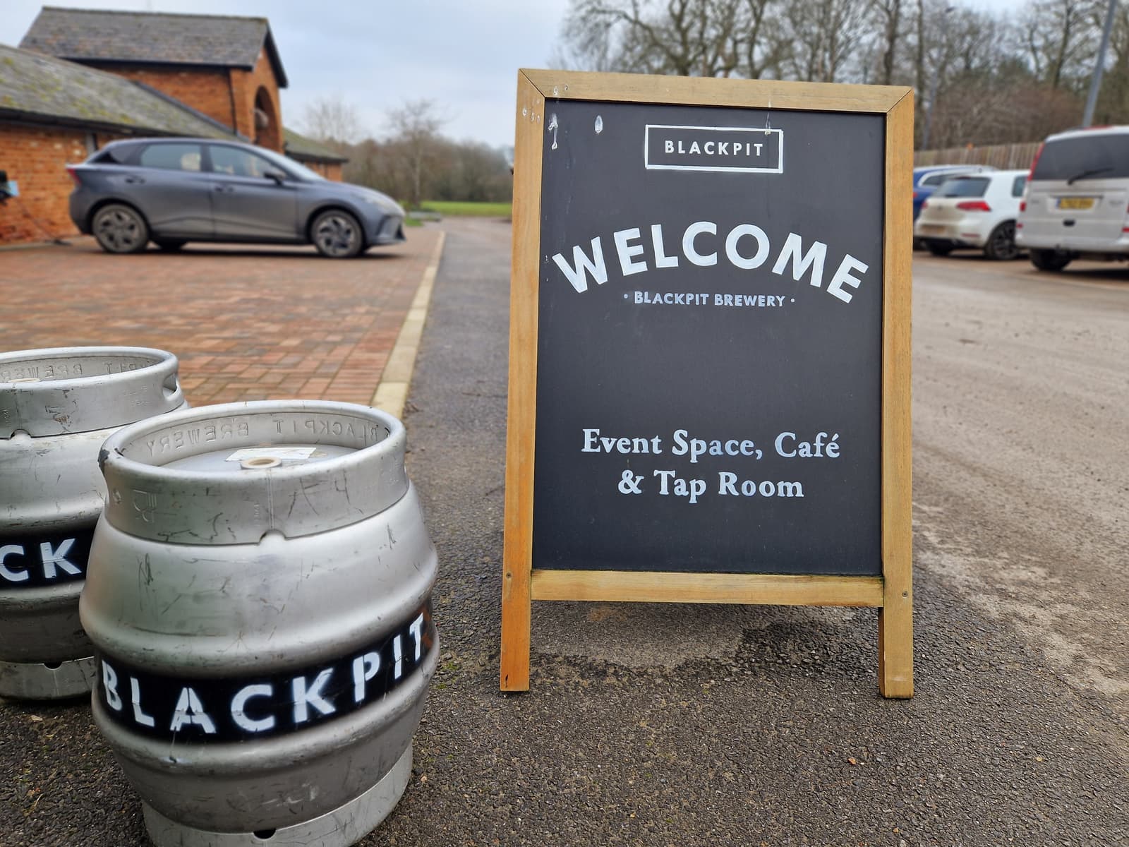 A chalkboard sign outside Blackpit Brewery reads "WELCOME, Event Space, Café & Tap Room." Two silver barrels marked "BLACKPIT" stand nearby, with cars and a red brick building in the background.