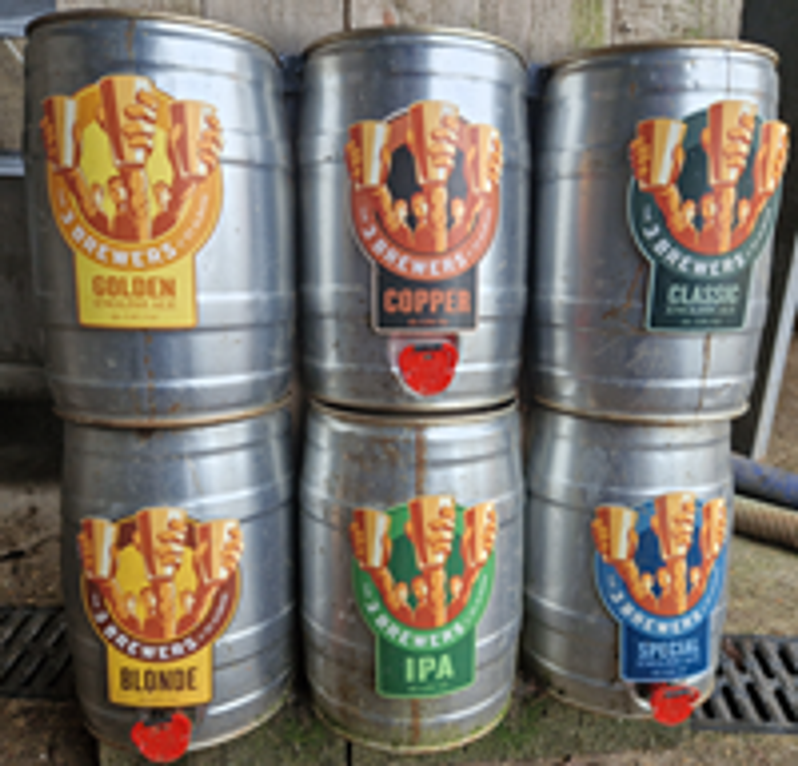 Six metal barrels stacked in two rows. Each barrel has a colorful label with a different beer type: Golden, Copper, Classic, Blonde, IPA, and Special. The labels feature a stylized logo with hands holding a barrel.