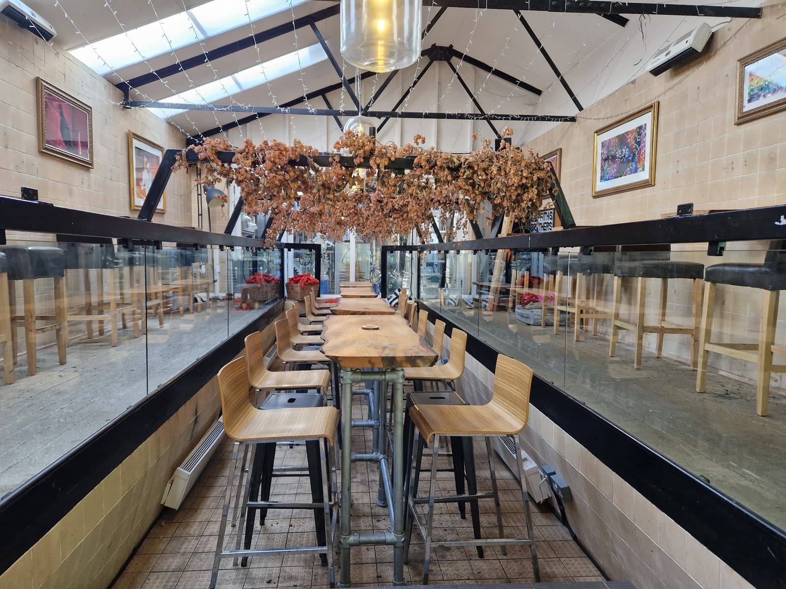 A long wooden table with stools runs down the center of a bright, glass-walled cafe. Dried leaves hang above, and framed art decorates the tan walls. Empty chairs and tables line the sides under a sloped ceiling.