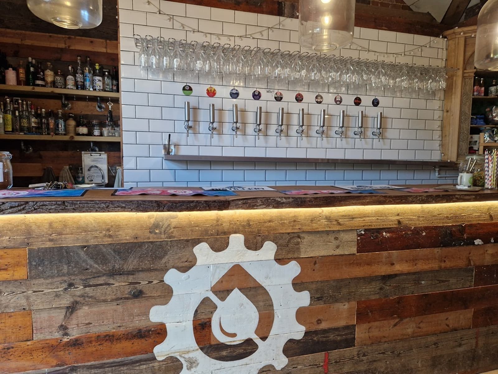 A rustic bar with a wooden counter displaying a gear and flame logo, taps for various beers, and shelves with bottles and glasses against a white subway tile wall.
