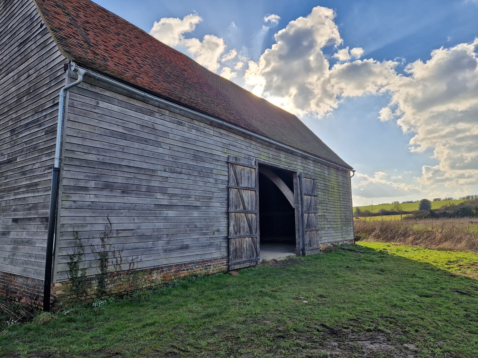 A wooden barn with open double doors sits on grassy ground under a partly cloudy sky, with sunlight peeking over the roof and a rural landscape in the background.