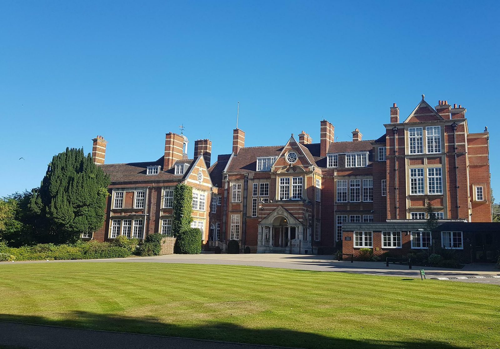 A large, historic building with a brick facade, multiple chimneys, and a symmetrical layout. The building is surrounded by manicured green lawns and some tall trees are visible to the left. The sky above is clear and blue.