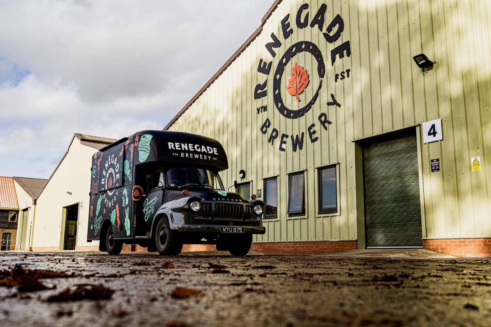 A vintage black truck with "Renegade Brewery" branding is parked in front of a large green warehouse building with "Renegade Brewery" signage. The ground is wet, reflecting the light, and there are scattered leaves. The building has a large garage door marked "4".