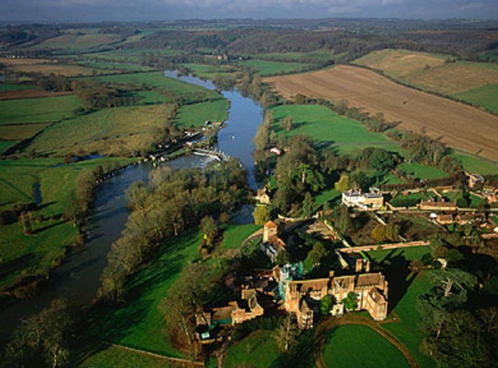 Aerial view of lush green landscape with a winding river flowing through farmland and woodlands. Several buildings and a large house with a garden are situated near the river. The terrain is a mix of fields, trees, and open countryside under a partly cloudy sky.