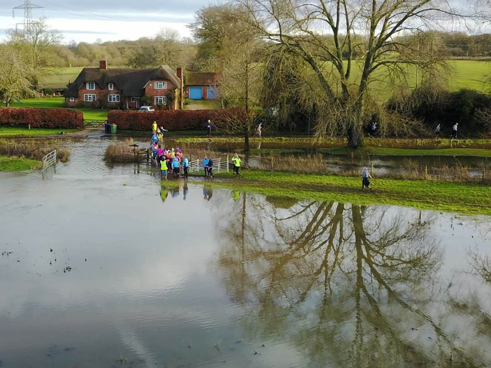 A large group of people cross a flooded path near a house with a thatched roof and trees reflected in the water, surrounded by green fields and bare trees. Some are running or walking on the grass beside the water. The sky is partly cloudy.