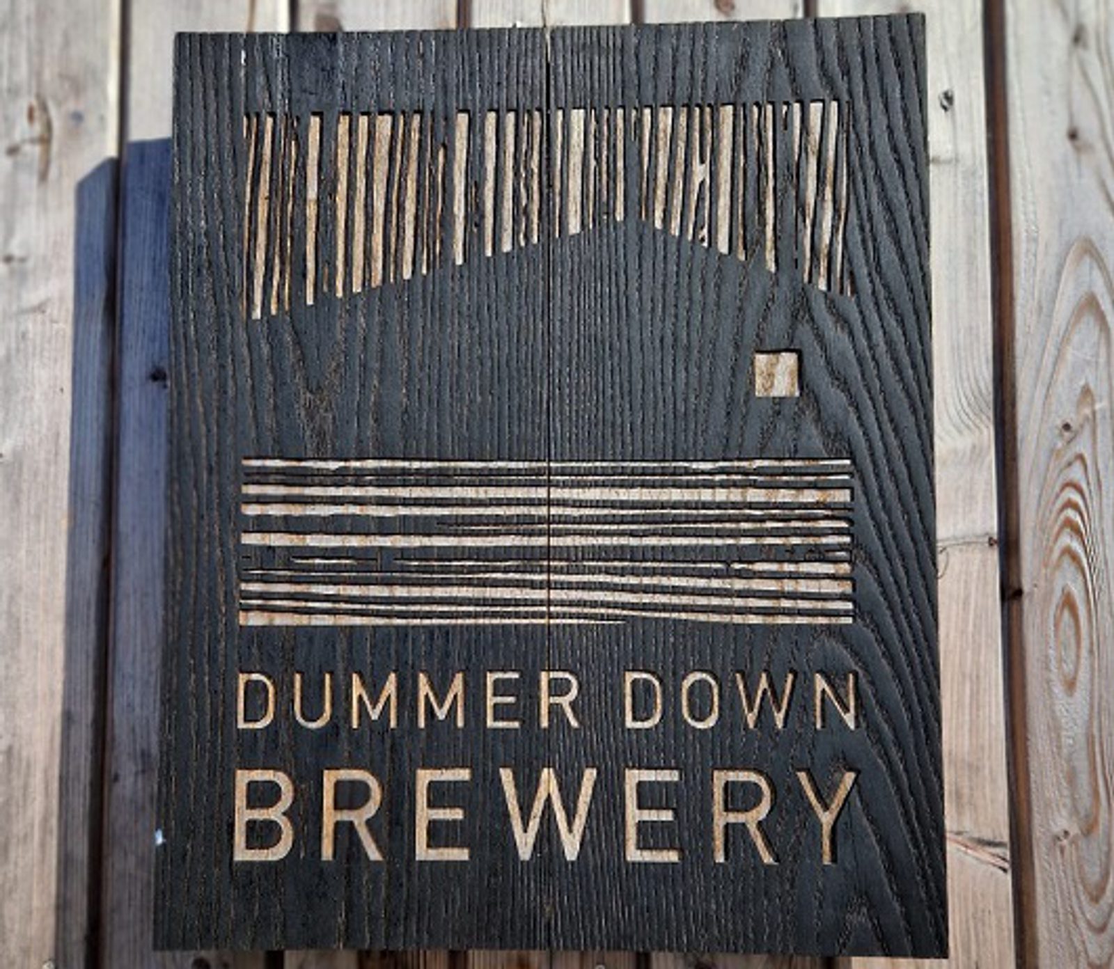 Wooden sign with a carved design of a house and lines, reading "Dummer Down Brewery," placed on a wooden background.