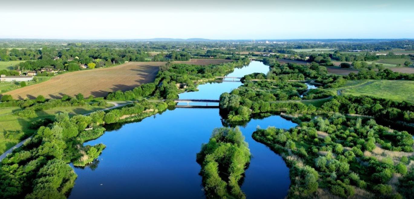 Aerial view of winding river surrounded by lush green trees and farmland. Several bridges span the river. The landscape extends into the distance with a mix of open fields, patches of forest, and scattered buildings under a clear blue sky.
