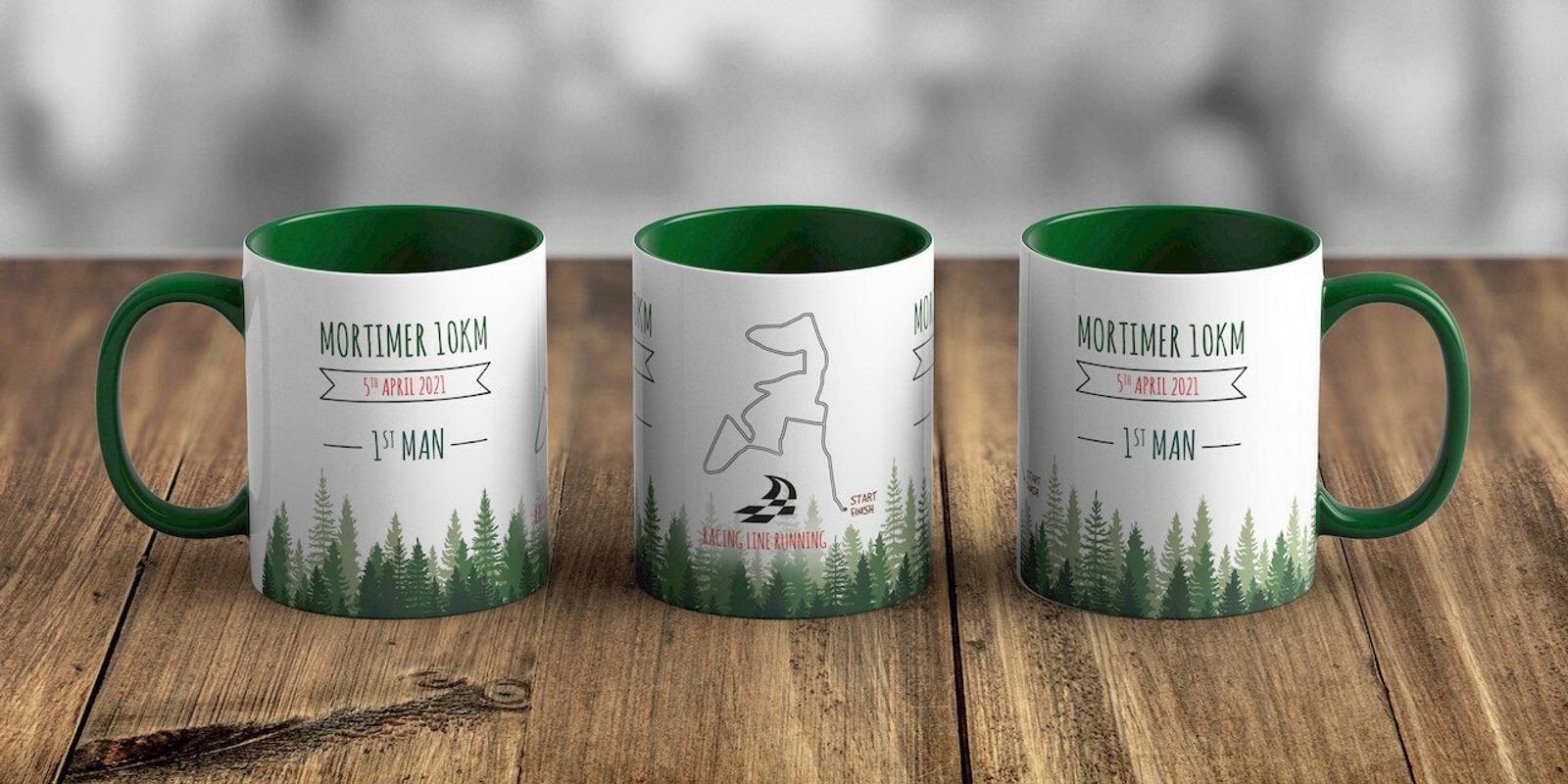 Three white and green mugs with a forest design and labeled "Mortimer 10KM, 9th April 2023, 1st MAN." The middle mug also features a runner silhouette and a logo with a running figure next to a sailboat. They are displayed on a wooden surface with a blurred background.