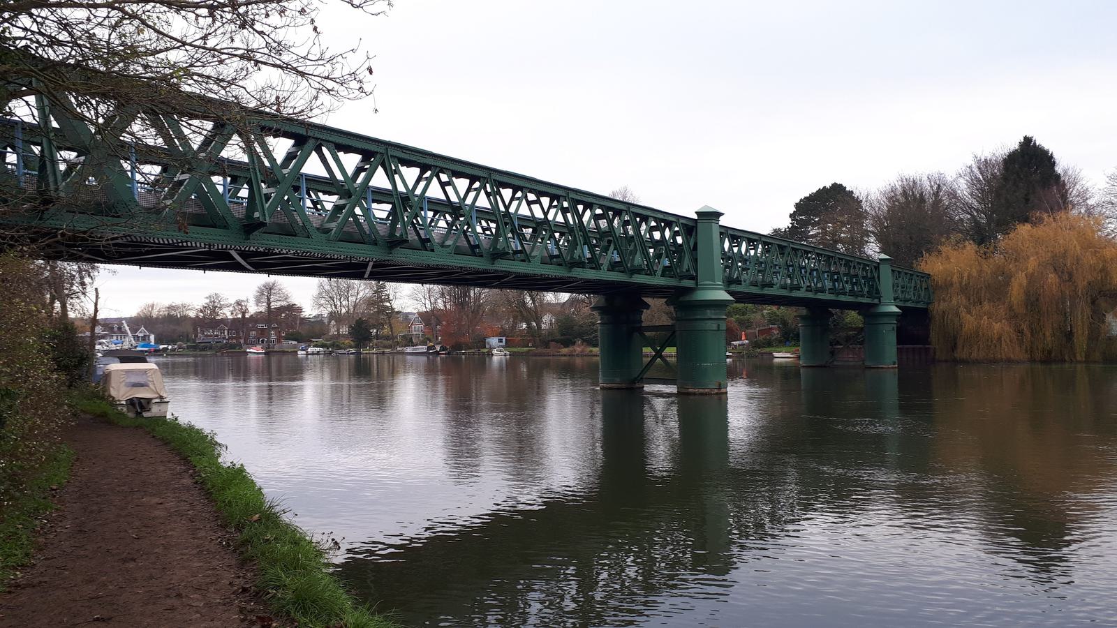 A green metal bridge spans over a calm river, with the water reflecting the structure. A boat is moored on the left bank, and a dirt path follows the river. Trees with autumn foliage line the banks, and buildings are visible in the background.