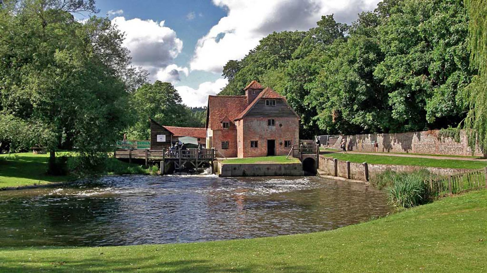 A picturesque scene of a historic water mill beside a gently flowing river. The mill is a brick building with a red-tiled roof, surrounded by lush greenery, trees, and a manicured lawn. A partly cloudy sky adds depth to the scenic countryside view.