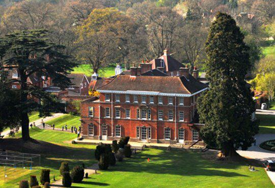 Aerial view of a large, historic red brick building surrounded by greenery and trees. The building has a symmetrical design with large windows, a central entrance, and a pitched roof. People are walking along the paths that encircle the building.