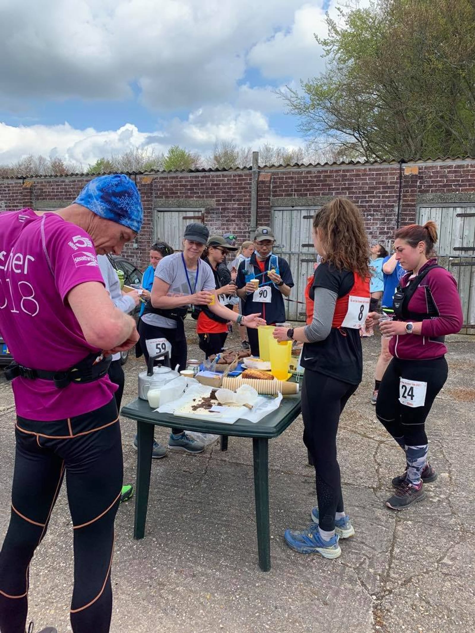 A group of runners gather around a table with refreshments during a race. They are wearing numbered bibs and athletic wear. Some are holding cups and eating snacks, while others look on. The setting appears to be outdoors on a cloudy day, with trees and a brick wall in the background.