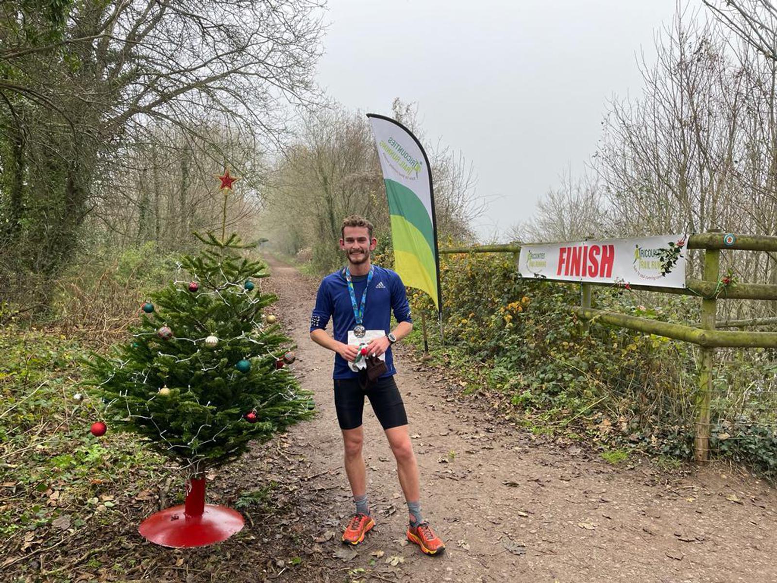 A man stands at the finish line of a trail race, holding a trophy and smiling. He is wearing a blue long-sleeve shirt, shorts, running shoes, and a medal around his neck. A decorated Christmas tree and a "FINISH" banner are beside him on a forest path.