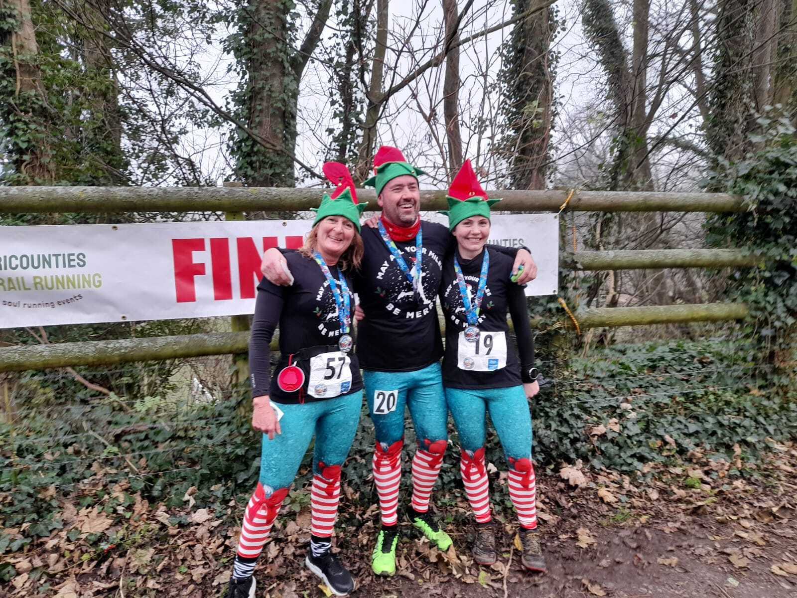 Three runners dressed in festive elf outfits, including red and green hats and matching leggings, celebrate at a trail running event finish line. They are wearing medals and have race numbers 57, 20, and 19. The backdrop includes trees and a "FINISH" banner.