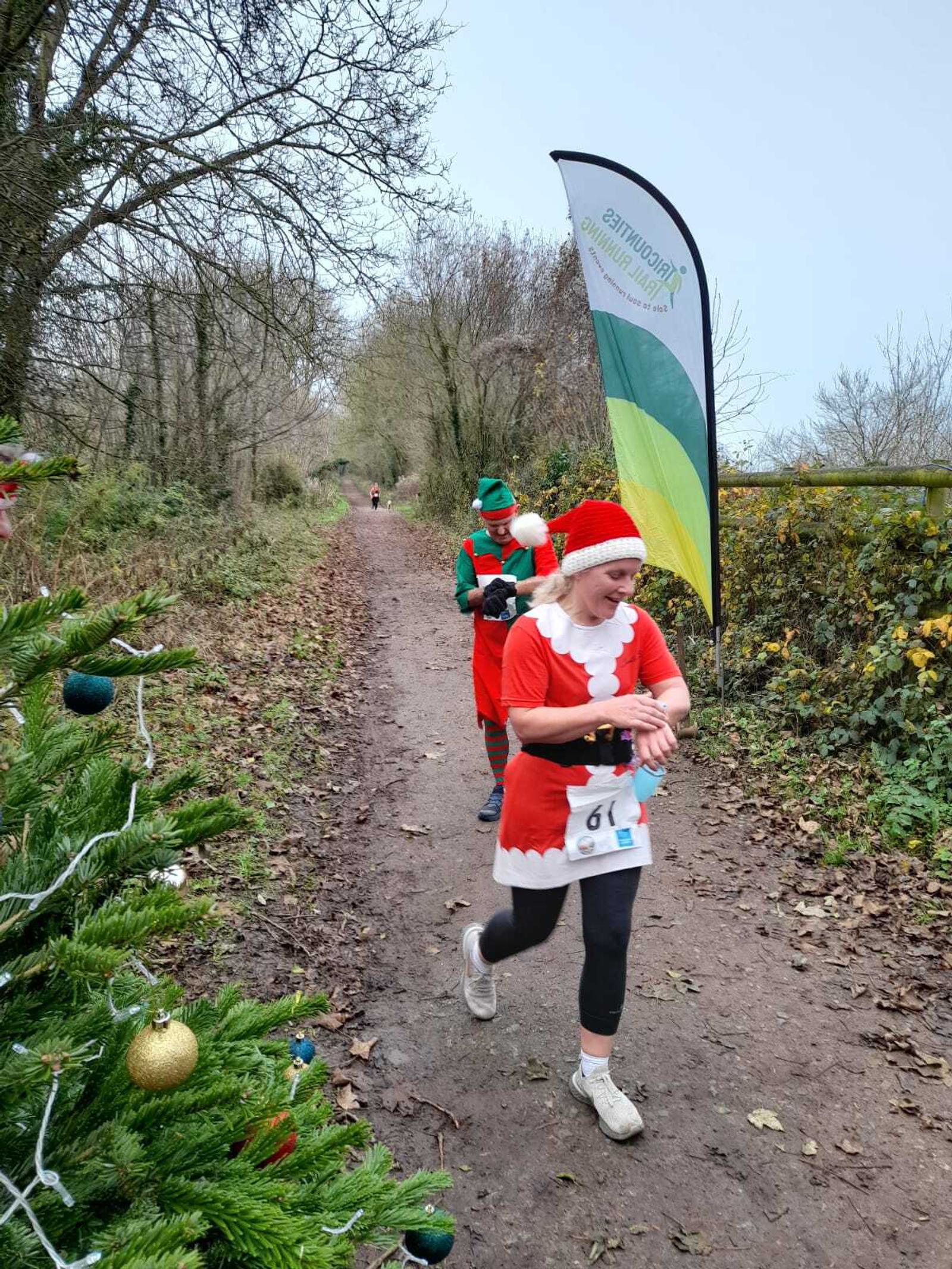 Two runners in festive costumes participate in a race through a forest trail. The foreground runner, dressed as Mrs. Claus, checks their watch. Behind them, another participant is dressed as an elf. A green and yellow banner is visible along the path.