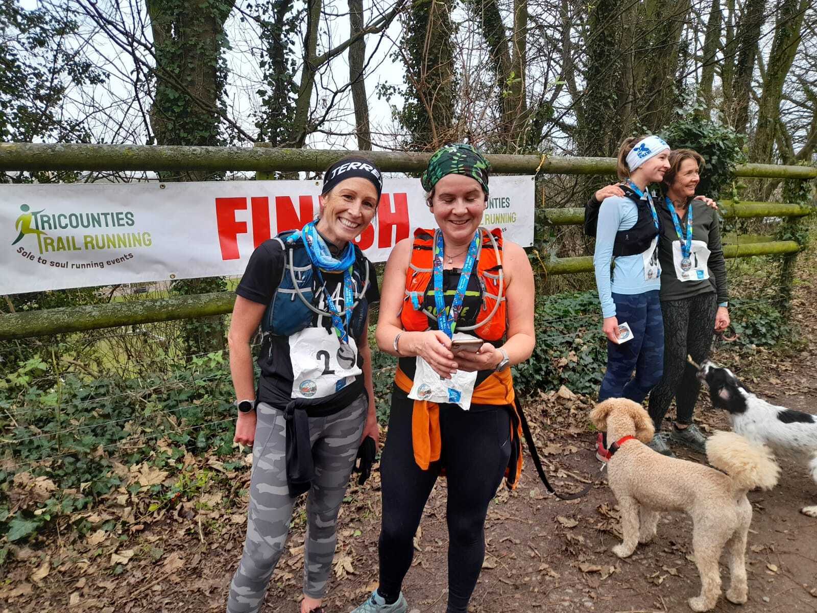 Two women in athletic gear smile at the camera after completing a race, standing in front of a "FINISH" banner. They are wearing medals and numbered bibs. Two dogs, one light brown and one black and white, stand nearby. Two other women are visible in the background.