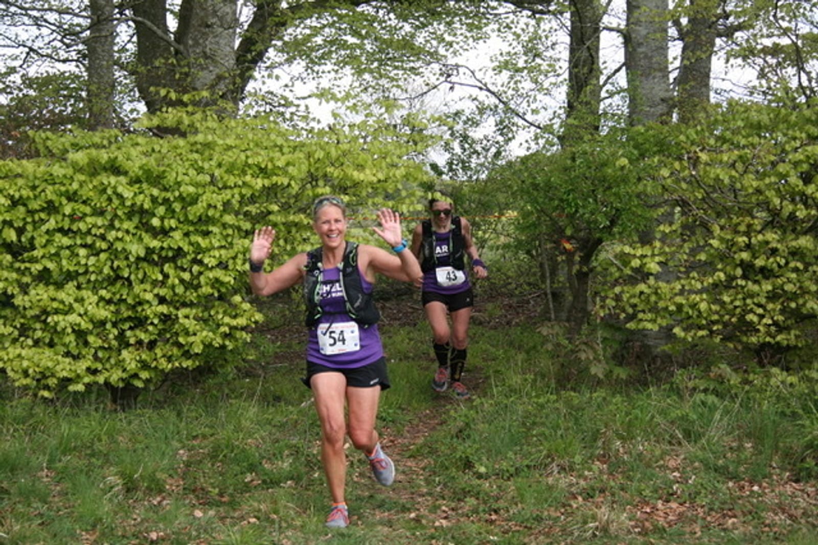 Two women are running on a grassy trail surrounded by trees and bushes. The woman in the foreground is smiling, wearing a purple tank top and numbered bib 54, and raising both hands. The woman behind her is also wearing a purple tank top and sunglasses.