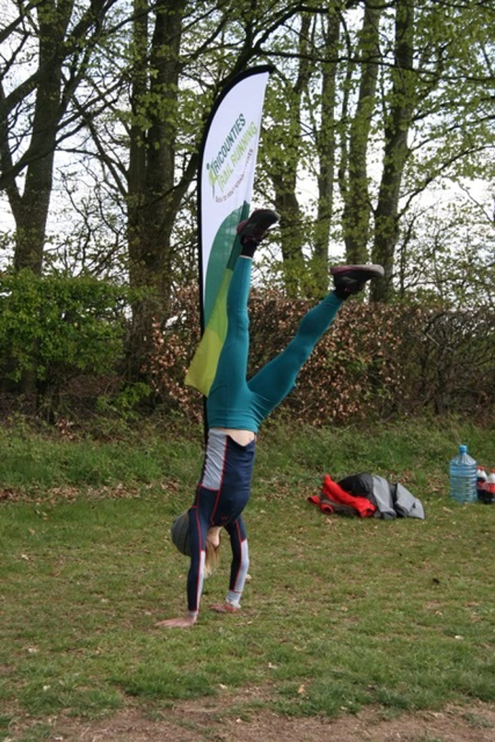 A person dressed in green and blue activewear is performing a handstand on a grassy area with trees in the background. There is a tall vertical banner with text and a logo, and various items like a water container and bags are on the ground nearby.
