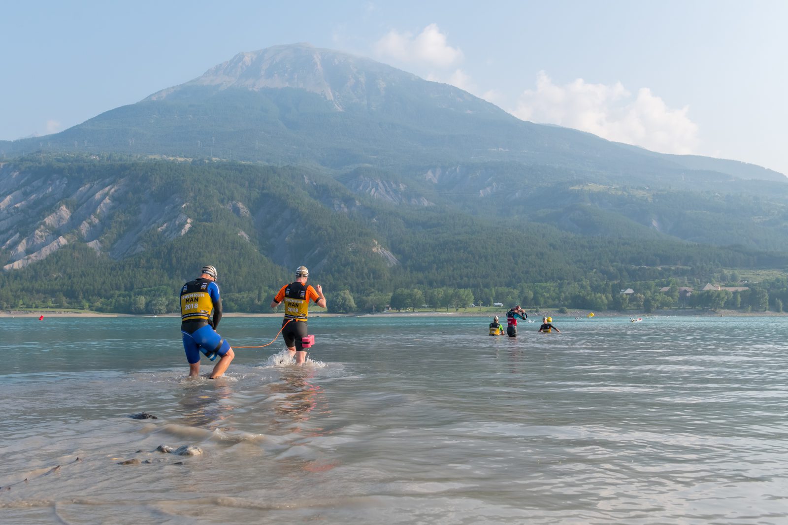 Athletes wearing wetsuits and swim caps wade through shallow water, heading towards deeper water for a swim competition. In the background, a large mountain and a lush green forested area can be seen under a clear sky.