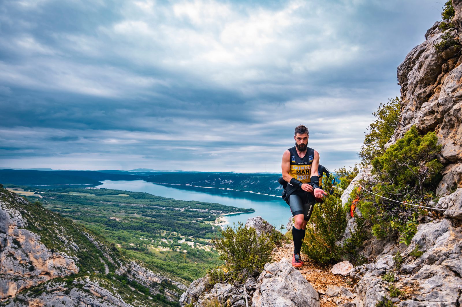 A bearded man trail running on a rocky mountain path with a lake and expansive greenery in the background. He is wearing a black and yellow athletic outfit and mid-length socks. The sky is overcast with dense clouds.