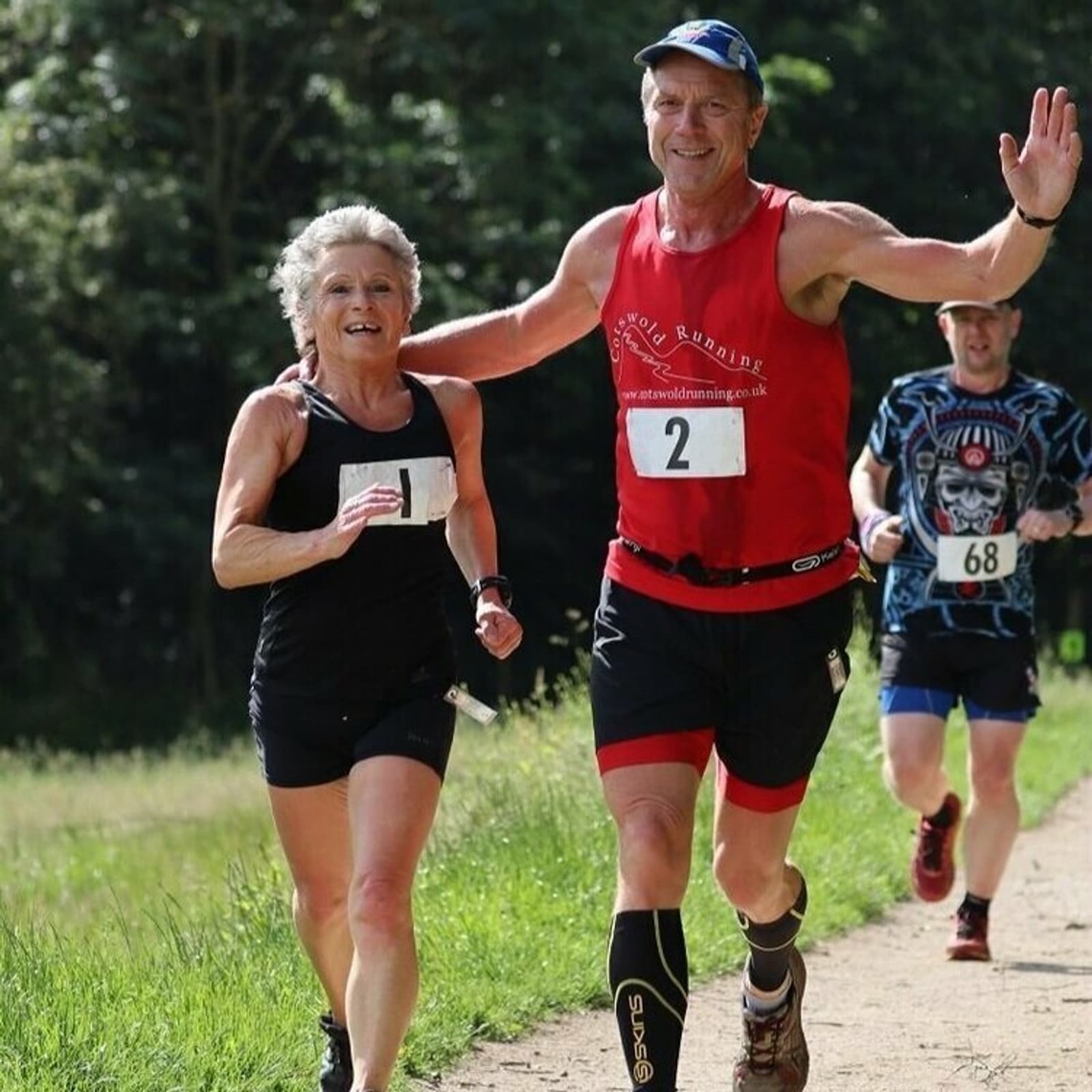 Three runners are on a sunny outdoor trail. One runner in a red tank top and black shorts, raising his arms, is beside another runner in a black tank top and shorts. In the background, a third runner in a blue patterned shirt and black shorts follows closely.