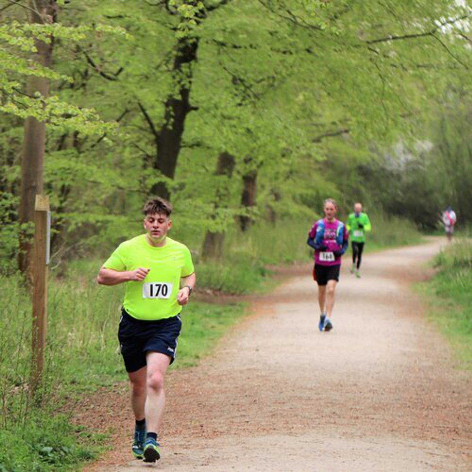 A young man wearing a neon yellow shirt and navy shorts runs on a dirt path surrounded by trees, with a race bib number 170. Behind him, two other runners are visible, one in a purple shirt and the other in green. The path is bordered by green foliage.