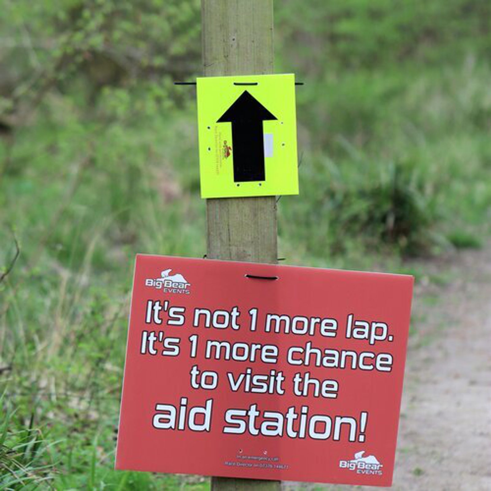A sign is posted on a wooden post along a trail. The top sign is yellow with a black upward arrow. Below it is a red sign with white text that reads, "It's not 1 more lap. It's 1 more chance to visit the aid station!" The background features greenery.