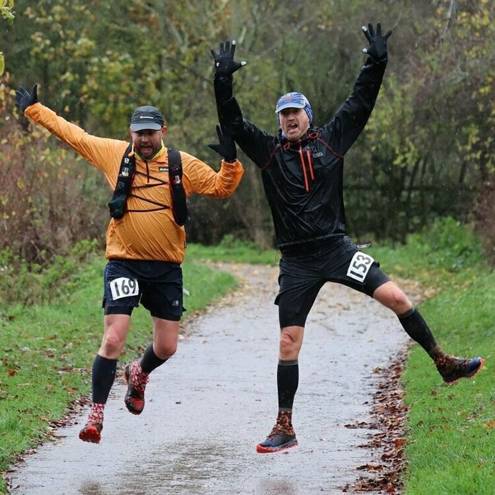 Two men wearing athletic gear are running exuberantly on a paved path through a park. Both have race bibs: one is 169 and the other 153. They are mid-jump with arms raised, surrounded by lush greenery and autumn foliage in the background.