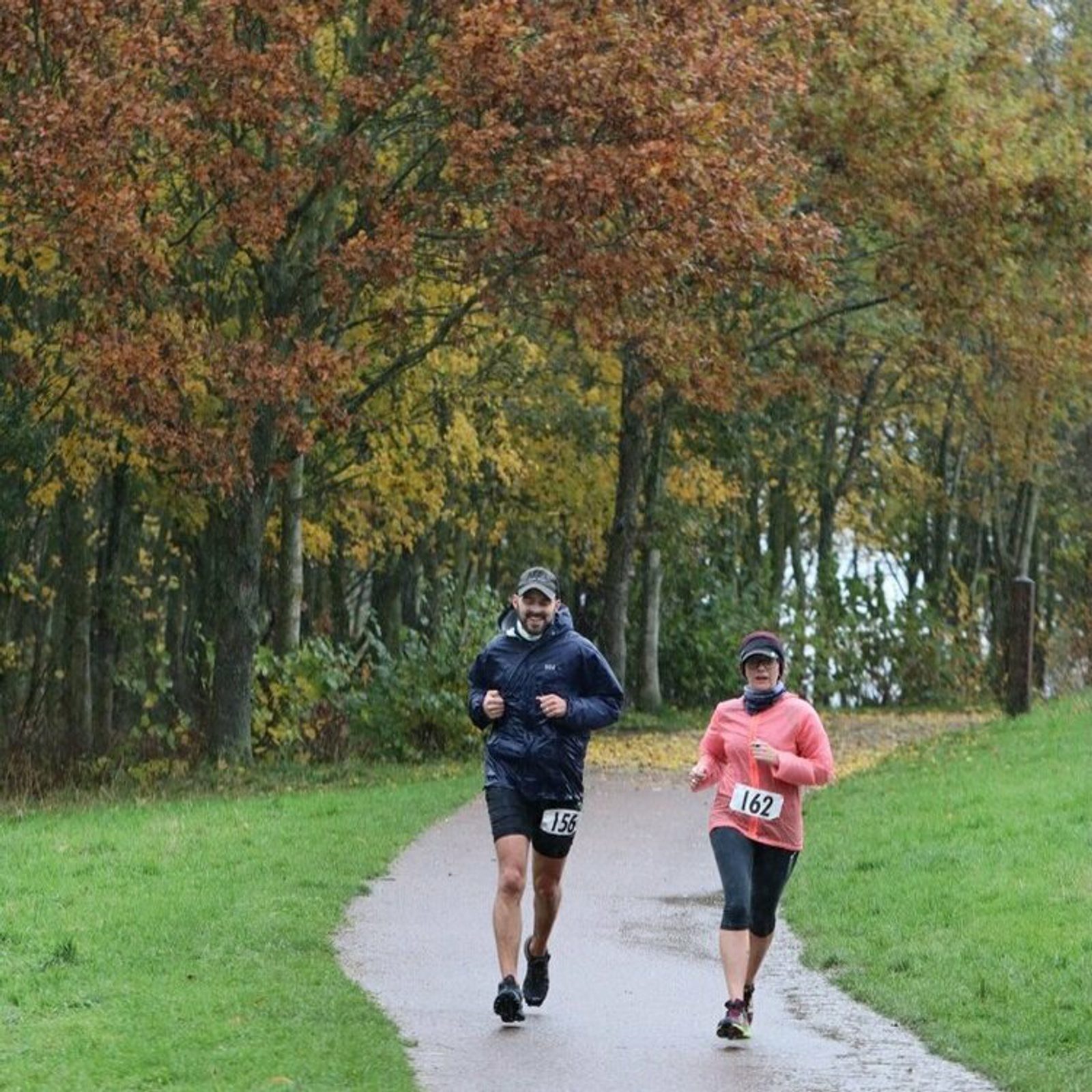 Two people, each wearing a numbered bib, jog on a path surrounded by autumn foliage. The person on the left wears black shorts and a dark jacket, while the person on the right wears a pink top and black capris. The ground is wet, suggesting recent rain.
