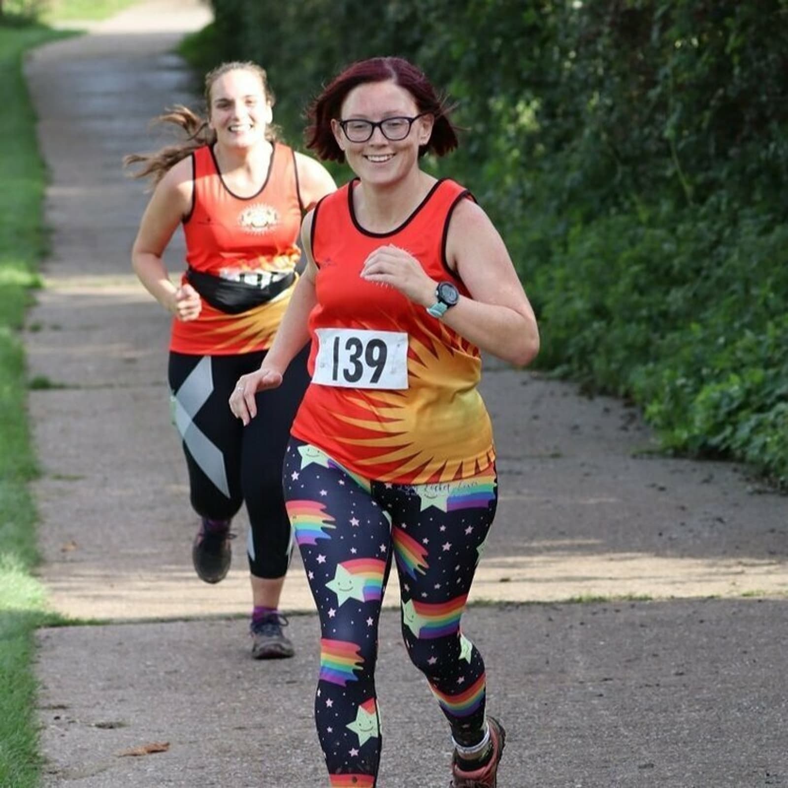 Two women are running on a paved path surrounded by greenery, smiling towards the camera. The woman in front is wearing a red and orange tank top with the number 139 and colorful leggings. The woman behind is wearing a similar tank top and black leggings.