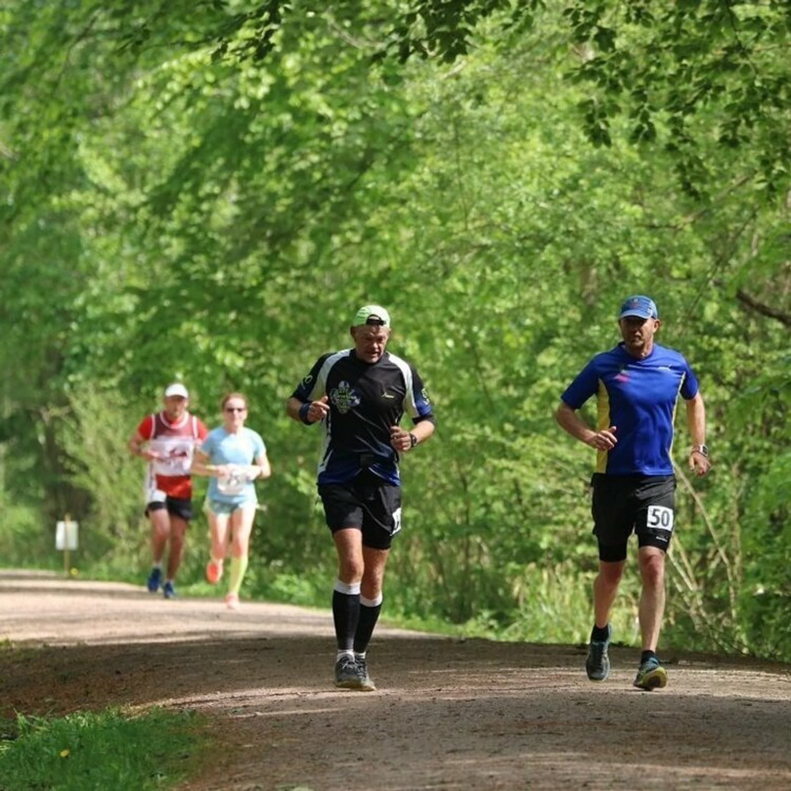 Four runners are running on a shaded forest trail. The leading runner wears a blue shirt and cap with the number '50' on their shorts, and is followed by a runner in a black outfit. Two more runners in red and white shirts are in the background.