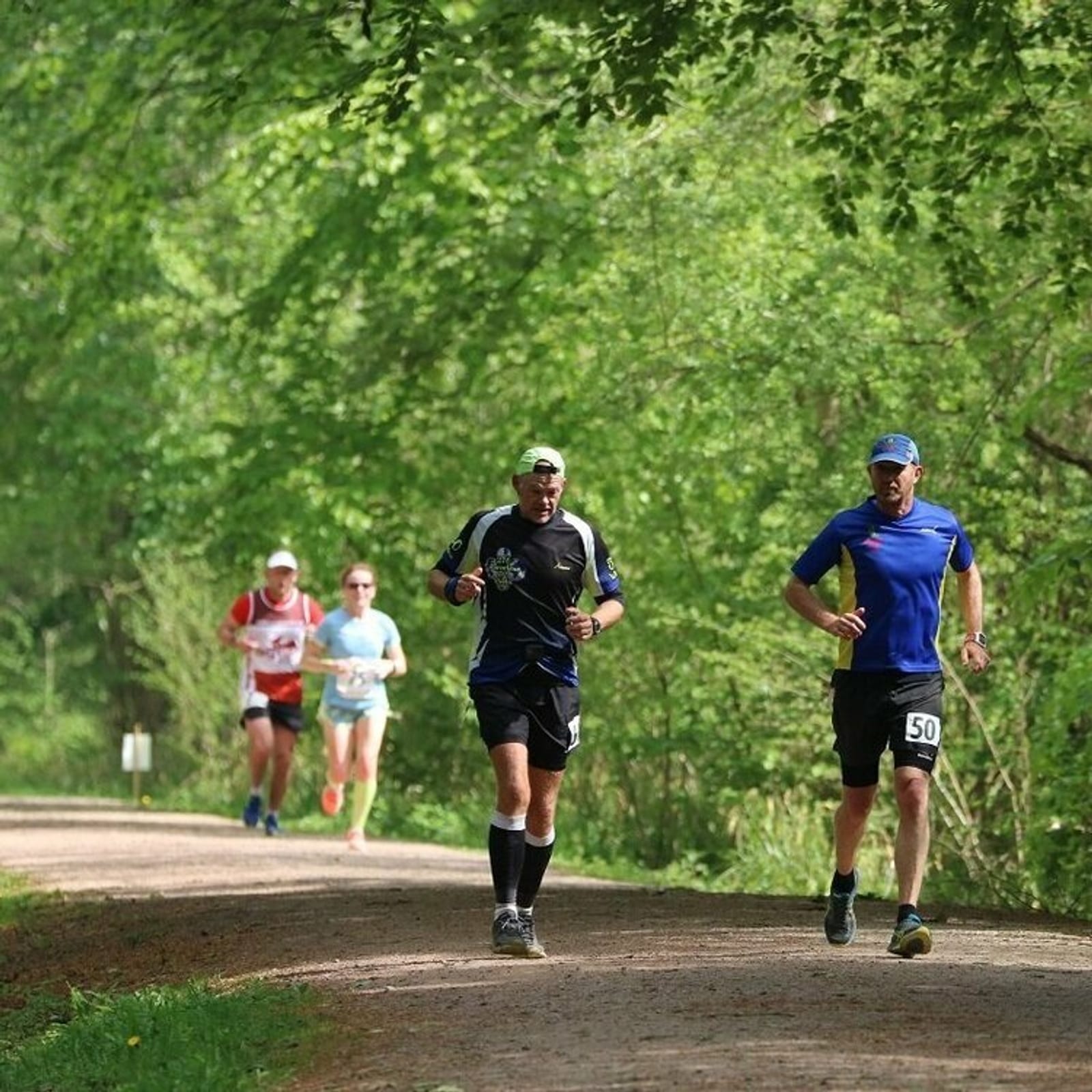 Four runners are participating in a race on a gravel path through a forested area. The path is surrounded by lush green trees, with two runners in the foreground and two in the background. The runner in front wears a cap and dark athletic gear, while the one behind them wears a blue shirt and shorts. The background runners are wearing red and light blue athletic outfits.