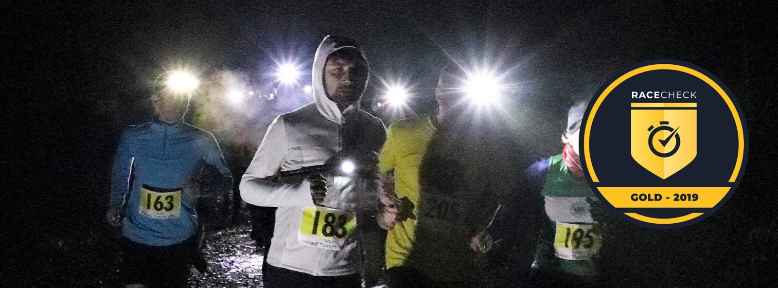 A group of runners, wearing headlamps and numbered bibs, participates in a nighttime race along a dark trail. The runner in the foreground wears a white hood and bib number 185. A "RACECHECK GOLD - 2019" badge is displayed on the right side of the image.