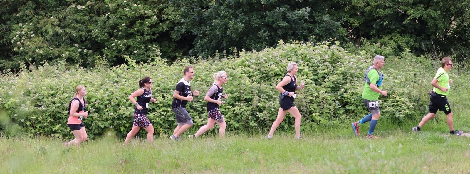 A group of six people is running outdoors on a grassy path. They are dressed in athletic clothing, some in black outfits and others in bright colors, with bib numbers attached. There are trees and bushes in the background under a cloudy sky.